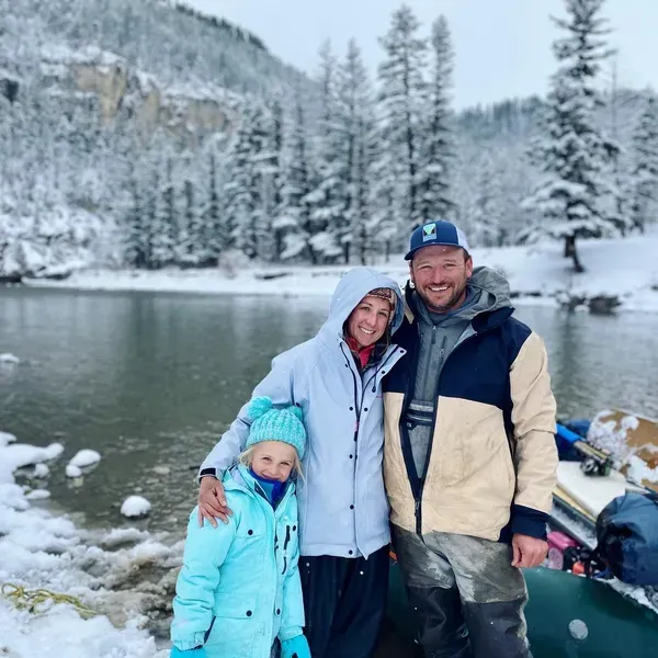 Family poses next to snowy river. Woman in light blue jacket, child in teal coat, man in jacket. Snow-covered trees in bg.