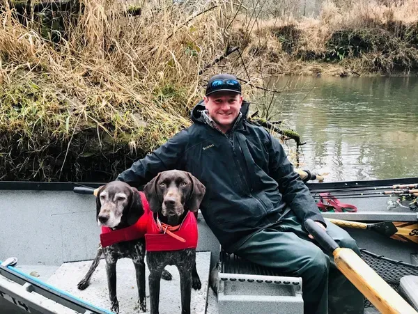 Man and two dogs in a boat on a river. Dogs wear red vests. Overcast day.