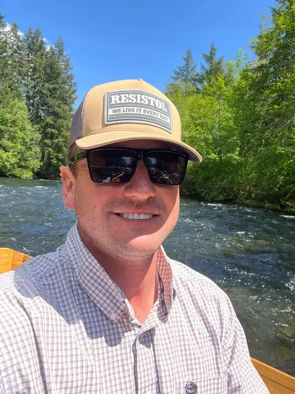 Man in tan hat and sunglasses smiles on a boat on a river, with trees in the background.