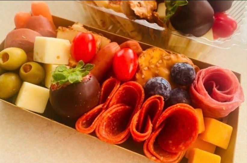 A cardboard box filled with a variety of food including cheese tomatoes olives and chocolate covered strawberries.