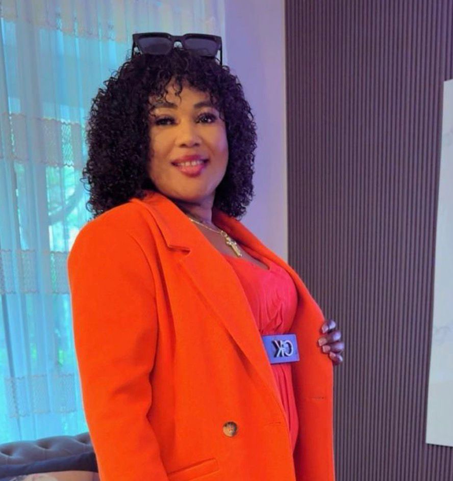 Eine Person mit lockigem Haar trägt einen  Blazer und posiert in einem Innenraum mit einer Sonnenbrille auf dem Kopf.