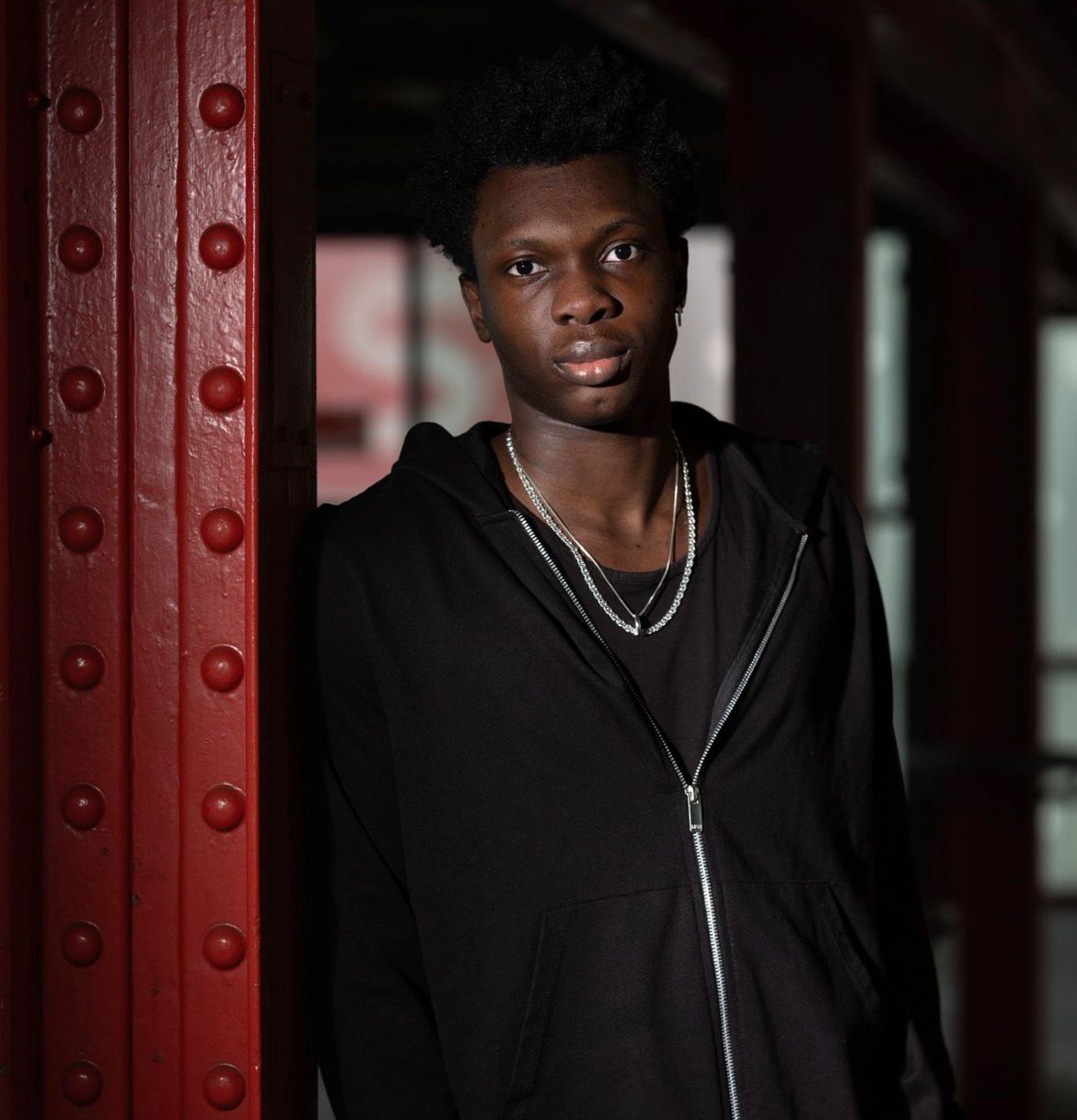 Eine Person mit dunklen Haaren, einen Kapuzenpullover mit Reißverschluss und Halsketten trägt, lehnt an einem Industrieträger