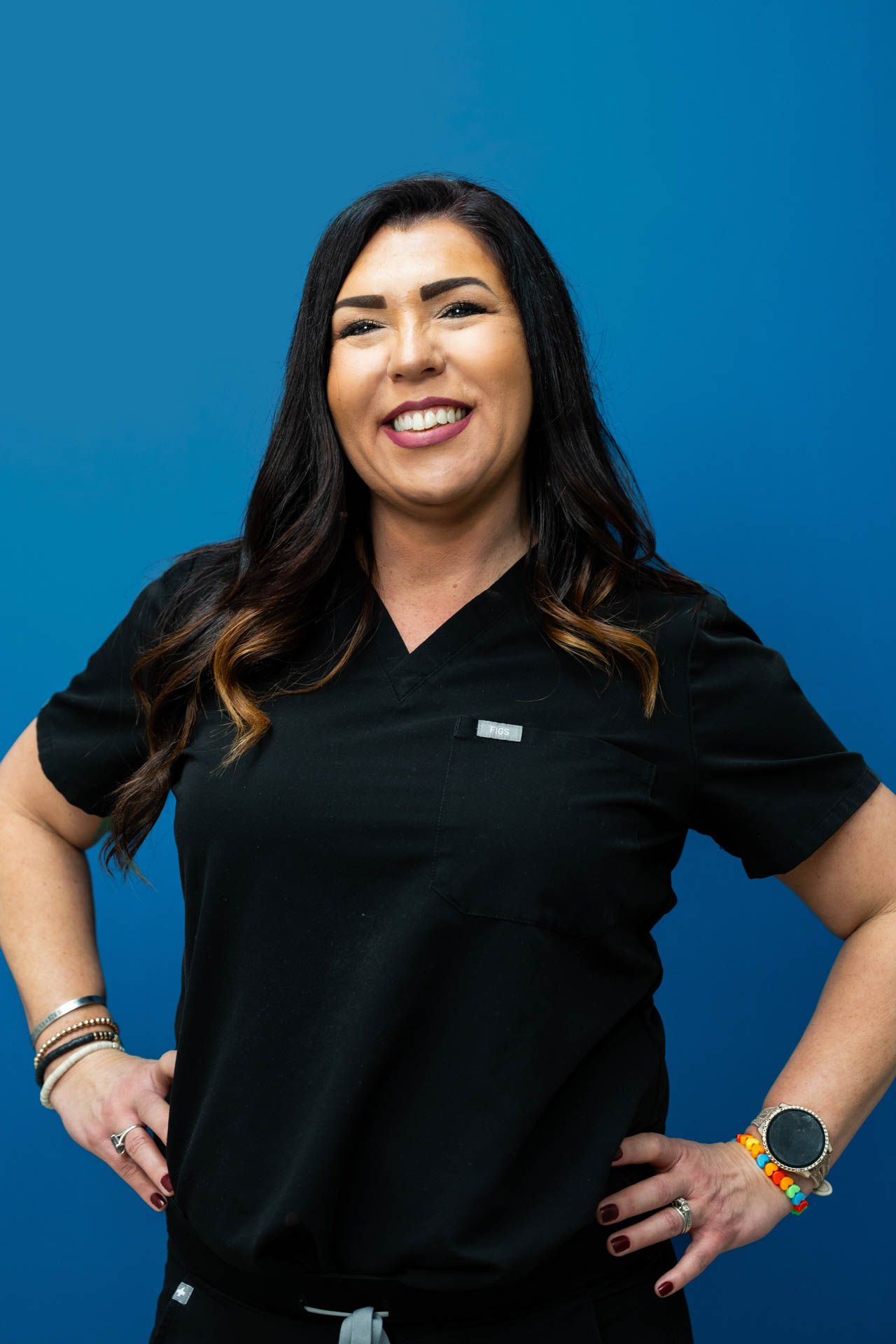 A woman in a black scrub top is standing with her hands on her hips and smiling.