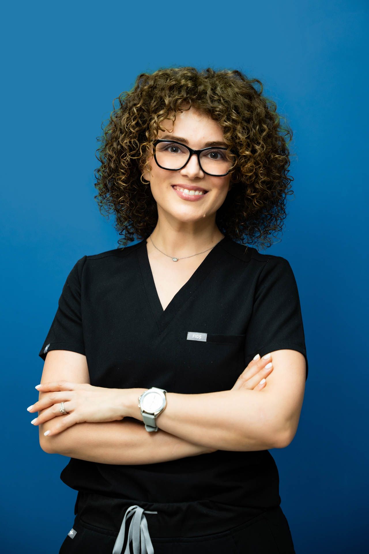 A woman wearing scrubs and glasses is standing with her arms crossed.
