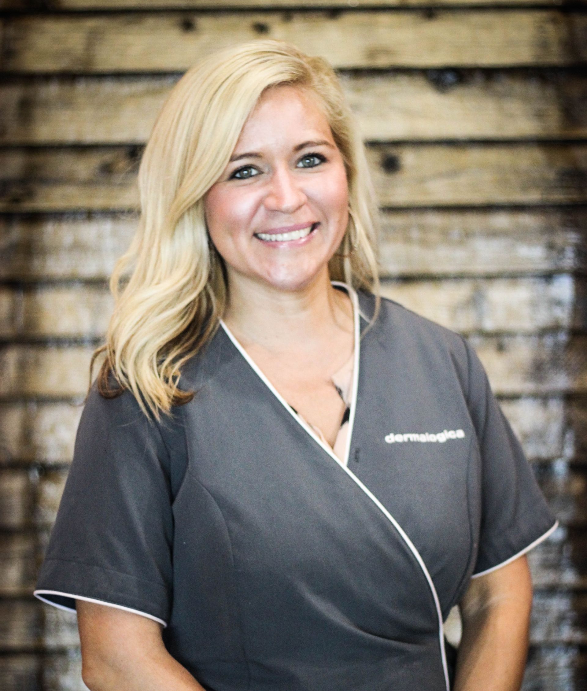 A woman in a grey scrub top is smiling in front of a wooden wall.
