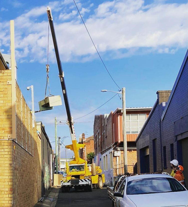 A Yellow Crane Is Lifting A Box In A Narrow Alleyway — Frigid-Air PTY Ltd In South Lismore, NSW