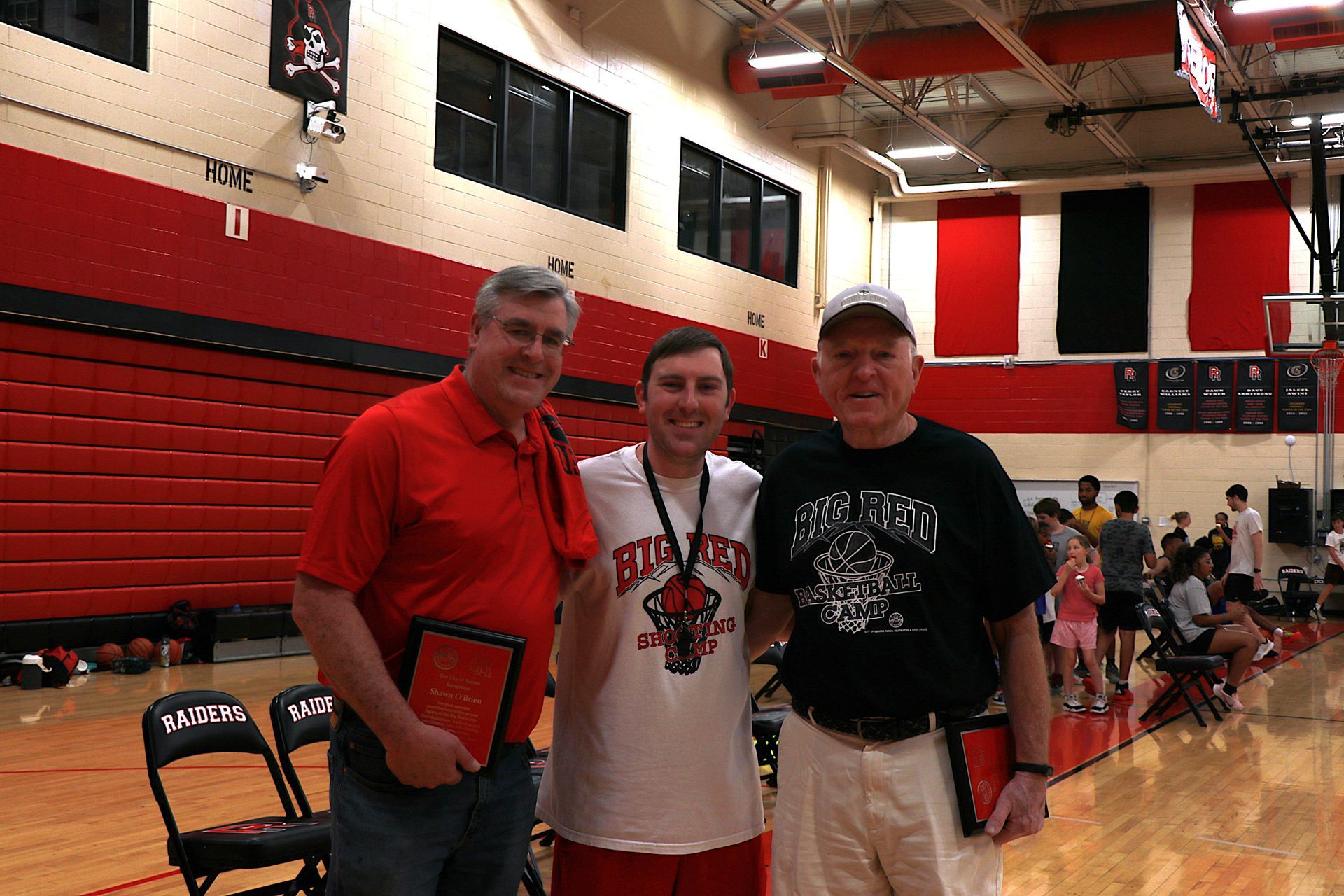 A person wearing a hat and orange shirt, holding a baseball bat on their shoulder on a baseball field.