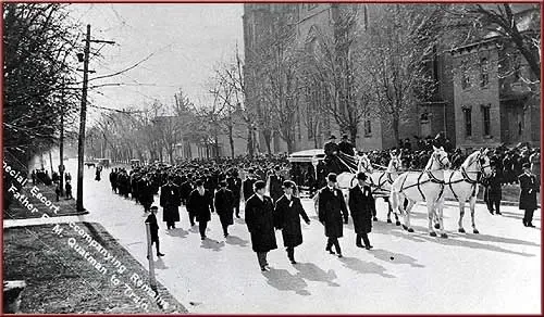 A black and white photo of a funeral procession with horse drawn carriages