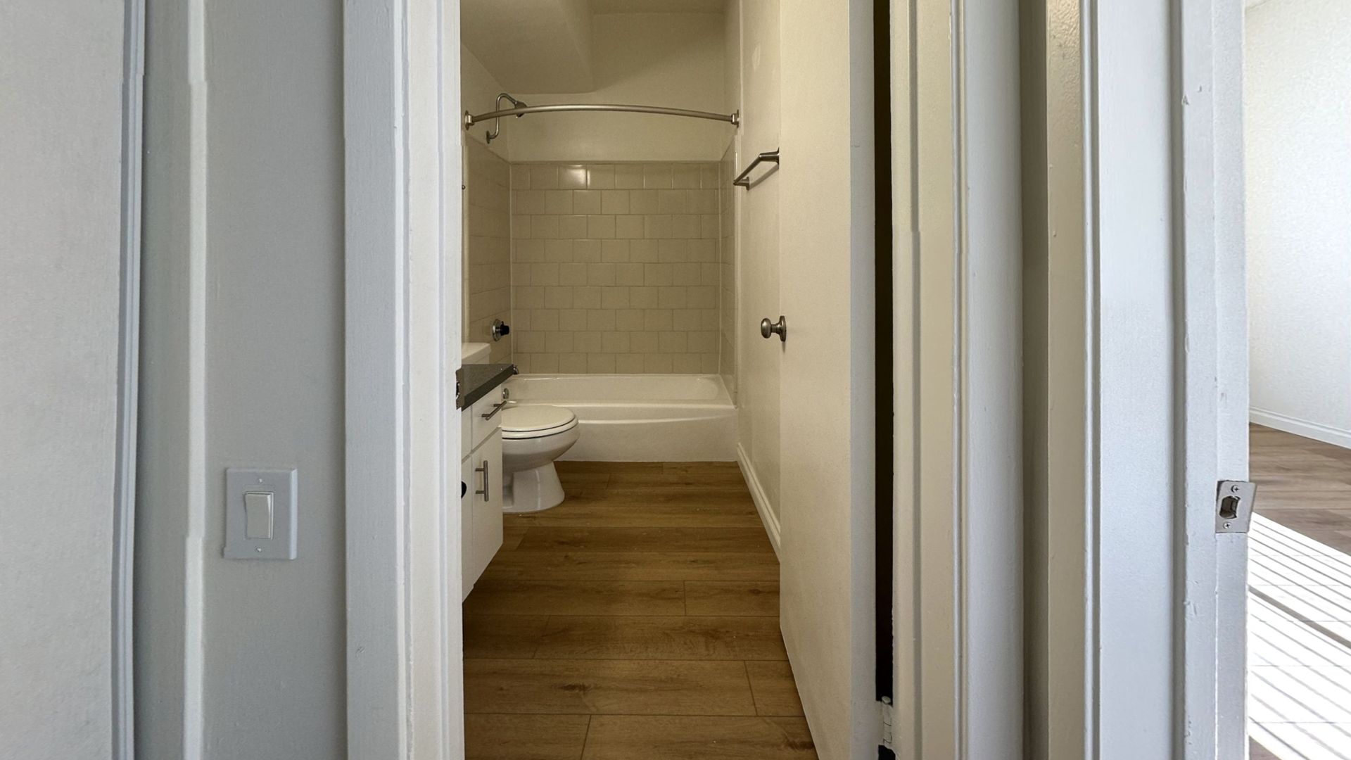 Hallway leading to a bathroom. Bathroom has a tub/shower, toilet, and sink. Wooden floor and white walls.