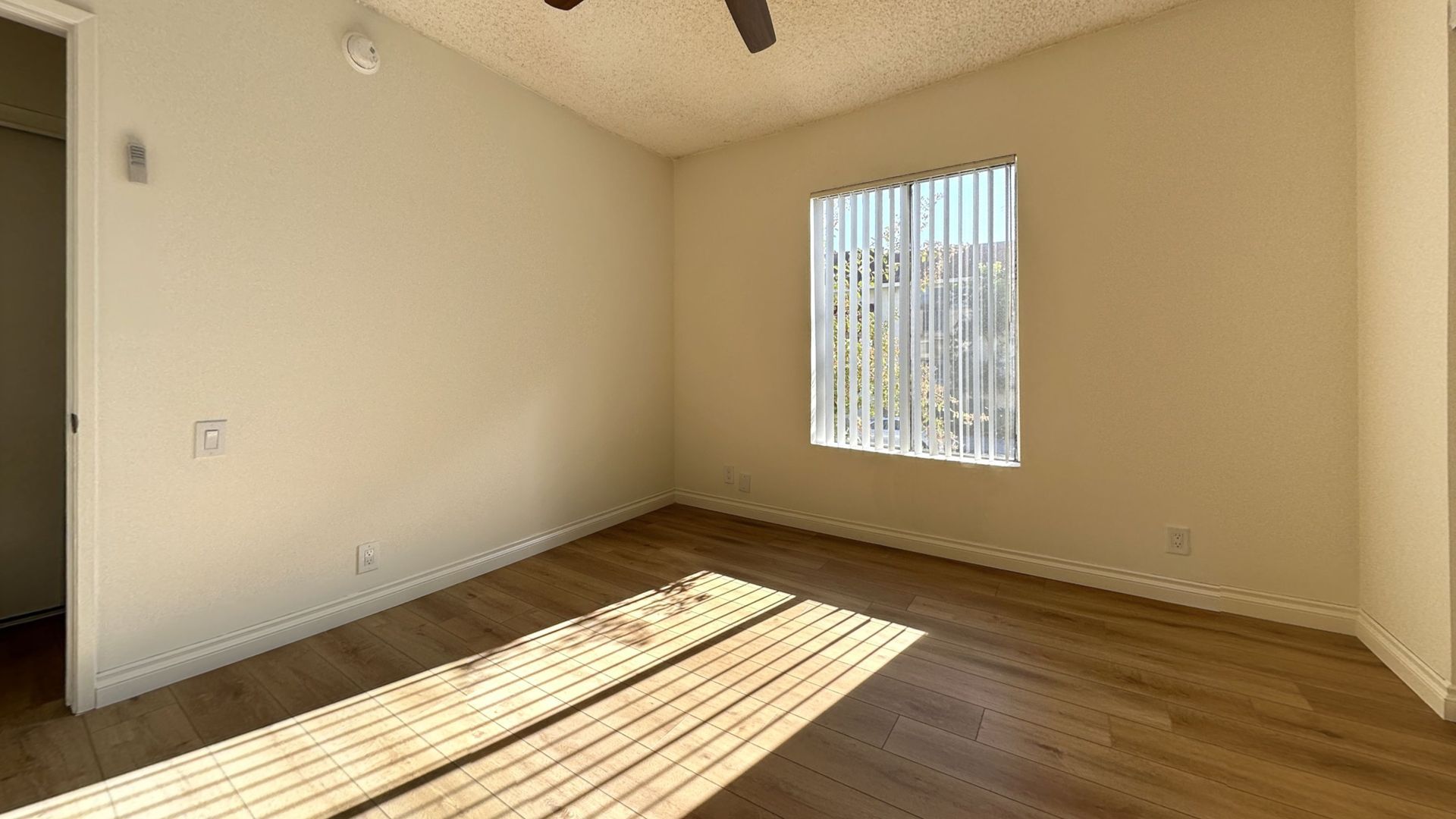 Empty room with wood floors and a window with vertical blinds, sunlit.