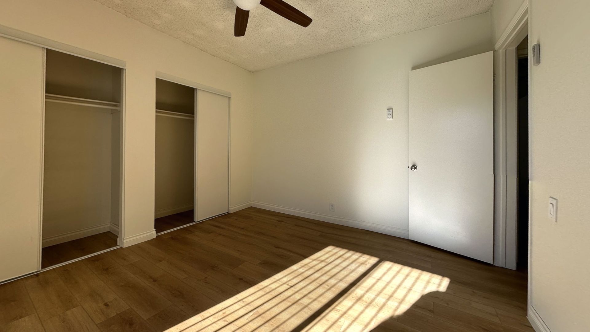 Empty bedroom with wooden floor, sliding closet doors, and a closed door. Sunlight streams through.