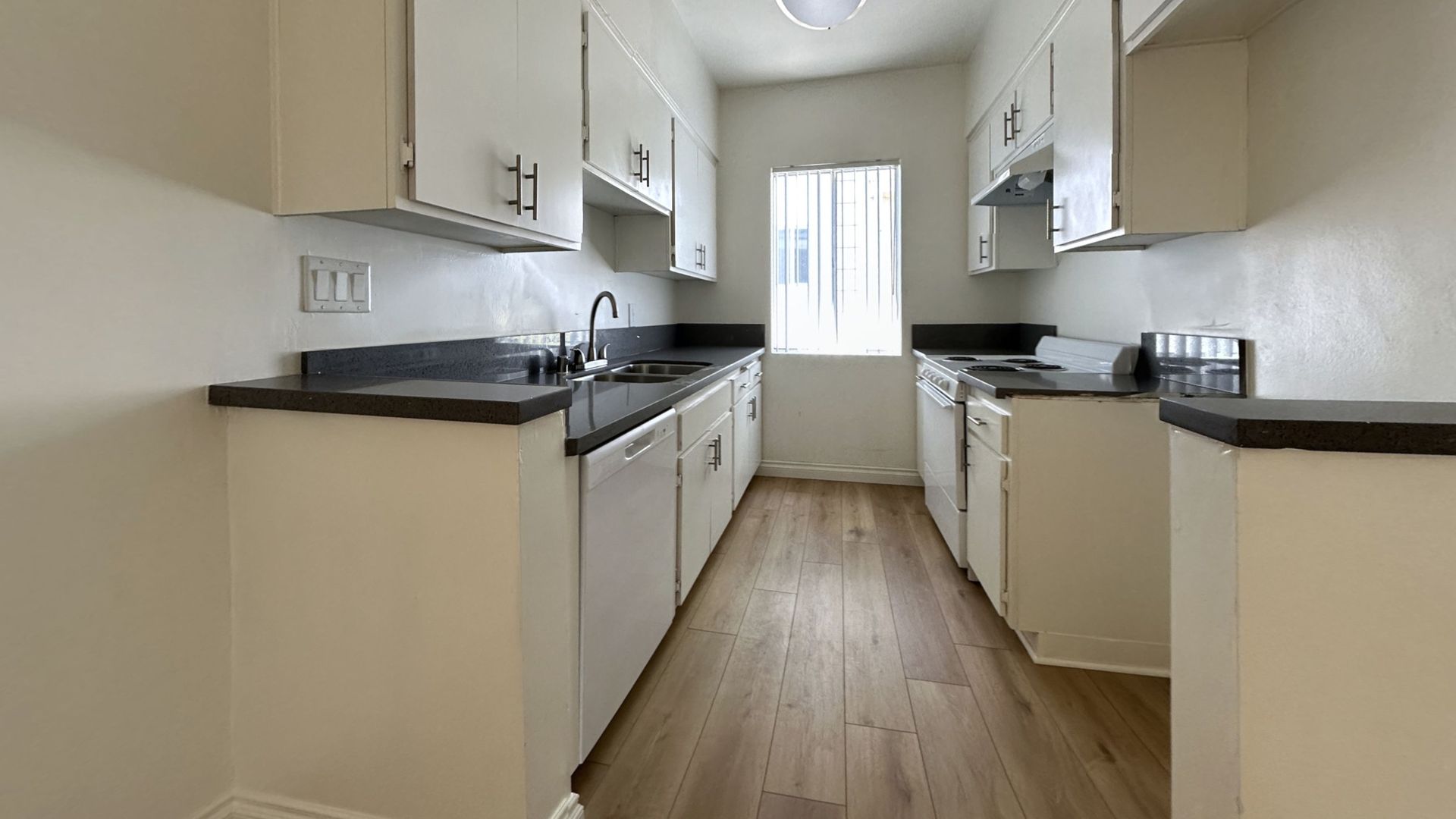 Narrow kitchen with white cabinets, dark countertops, and a window at the end.