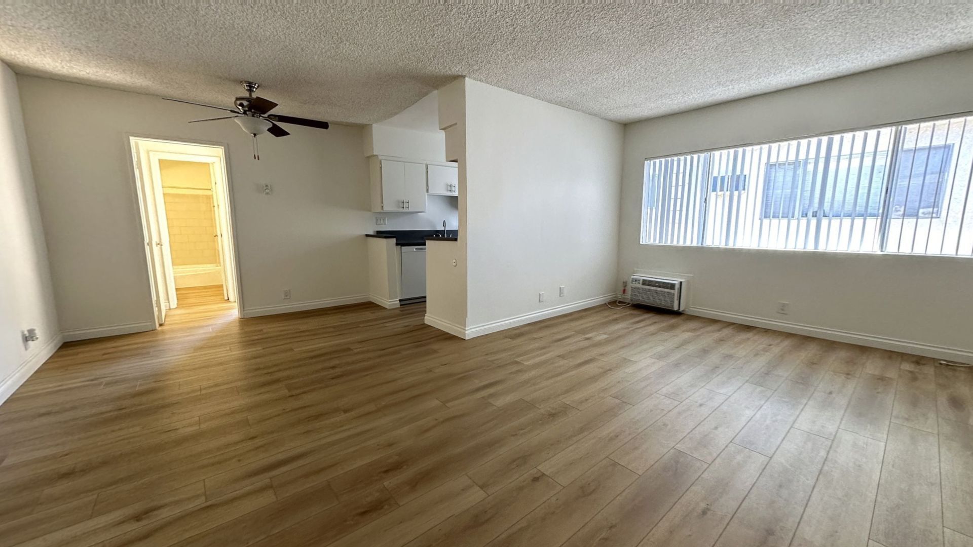 Empty apartment interior with wood flooring, kitchen, and bathroom visible.