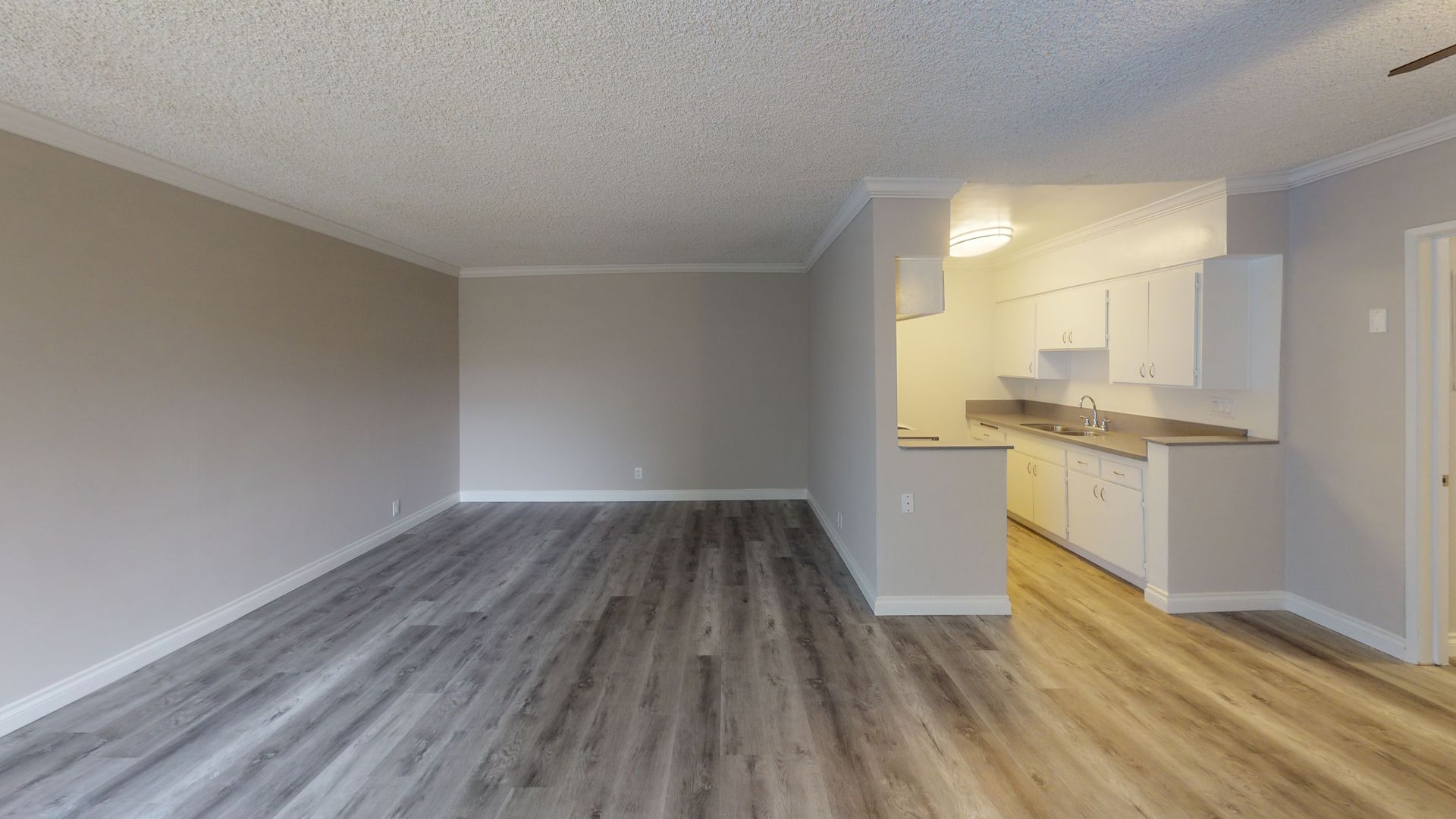 Empty apartment interior with wood-look flooring, light gray walls, and a small kitchen visible in the background.