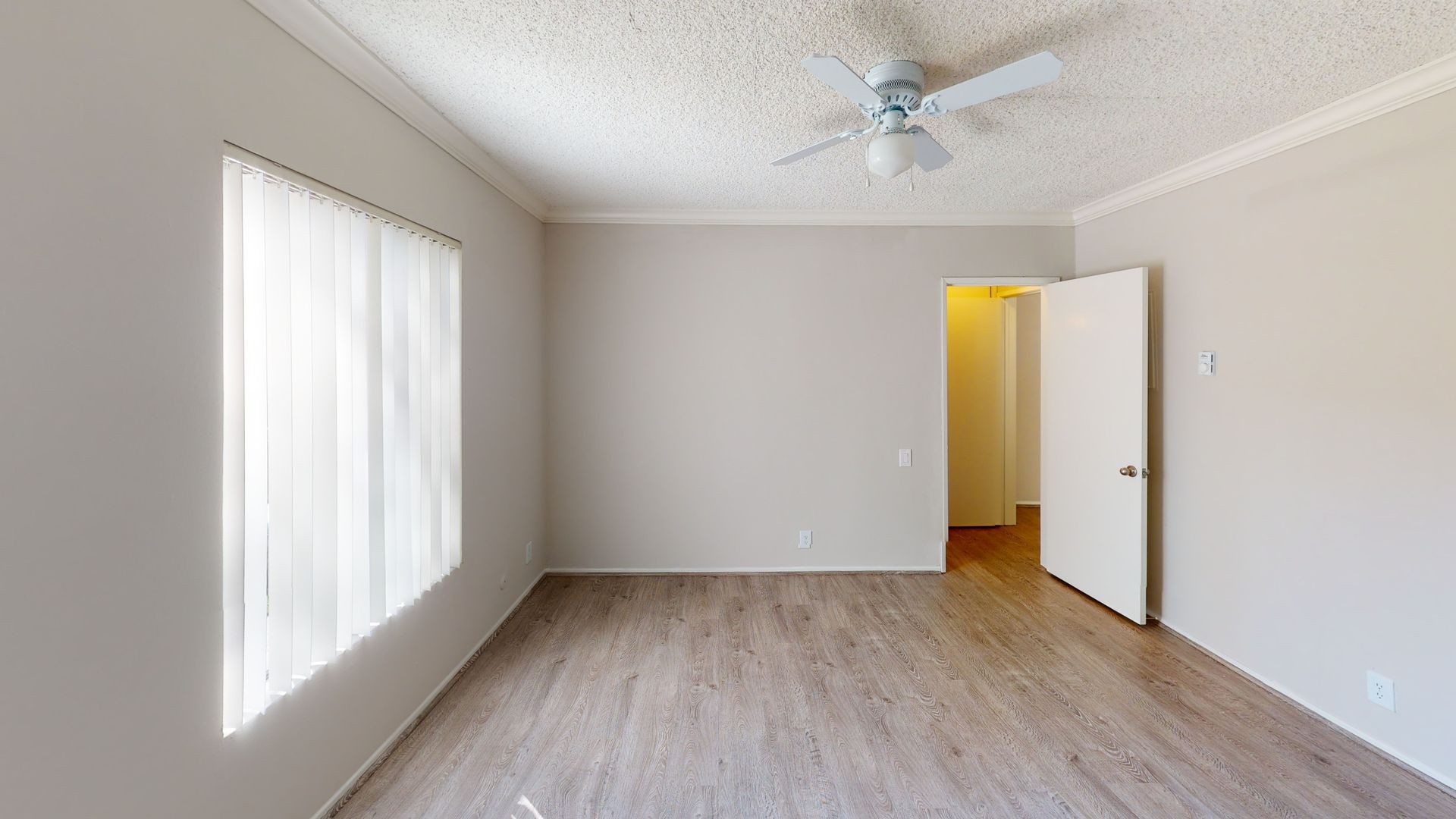 Empty room with light-colored walls, wood-look floor, window with sheer curtains, ceiling fan, and open door.