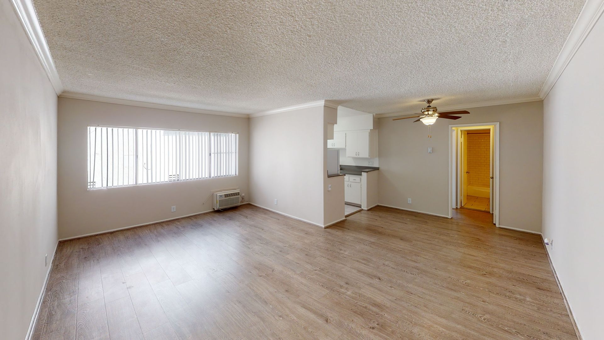 Empty apartment living room with wood-look flooring, white walls, textured ceiling, and a kitchen area.