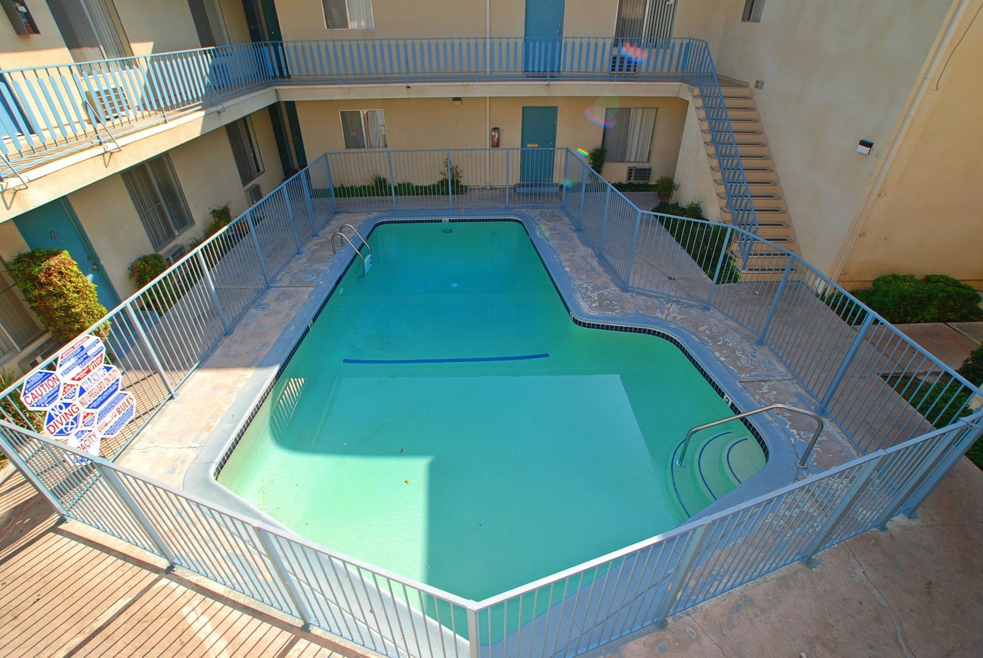 Swimming pool in a courtyard, surrounded by a light blue fence and apartment buildings.