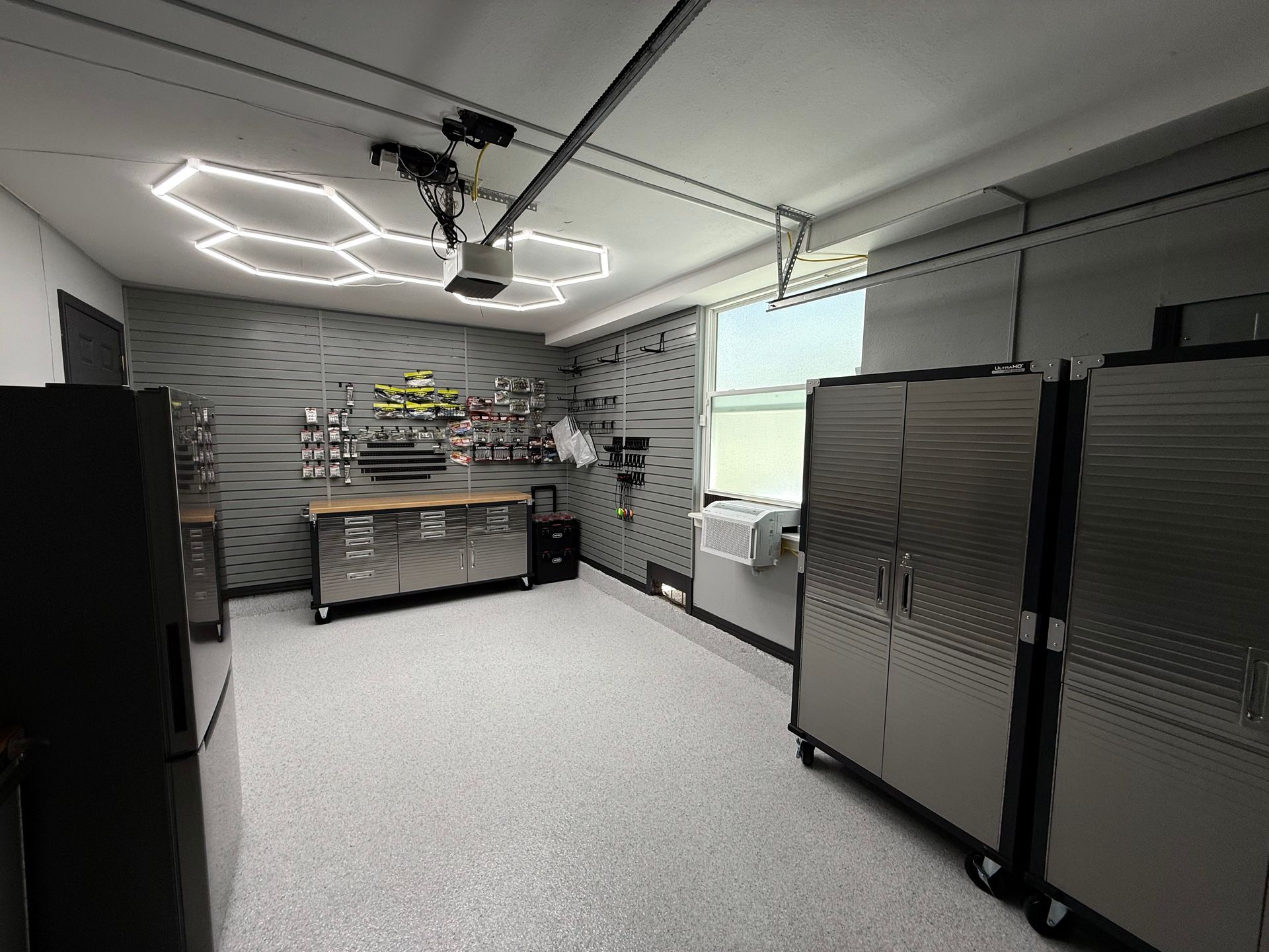 A well-lit garage with stainless steel cabinets, workbench, tools, and a refrigerator.