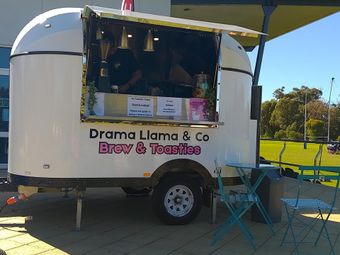 White coffee trailer labeled “Drama Llama & Co Brew & Toasties” parked on grass with outdoor table and chairs