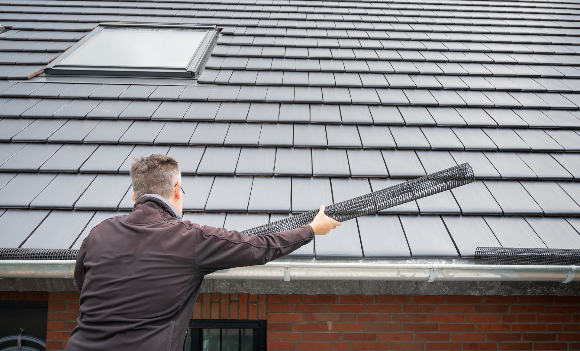 Man Installing Gutter Guard on a Tiled Roof. Gray Tiles, Brick Building — Callaway Roofing & Construction in Dubbo, NSW
