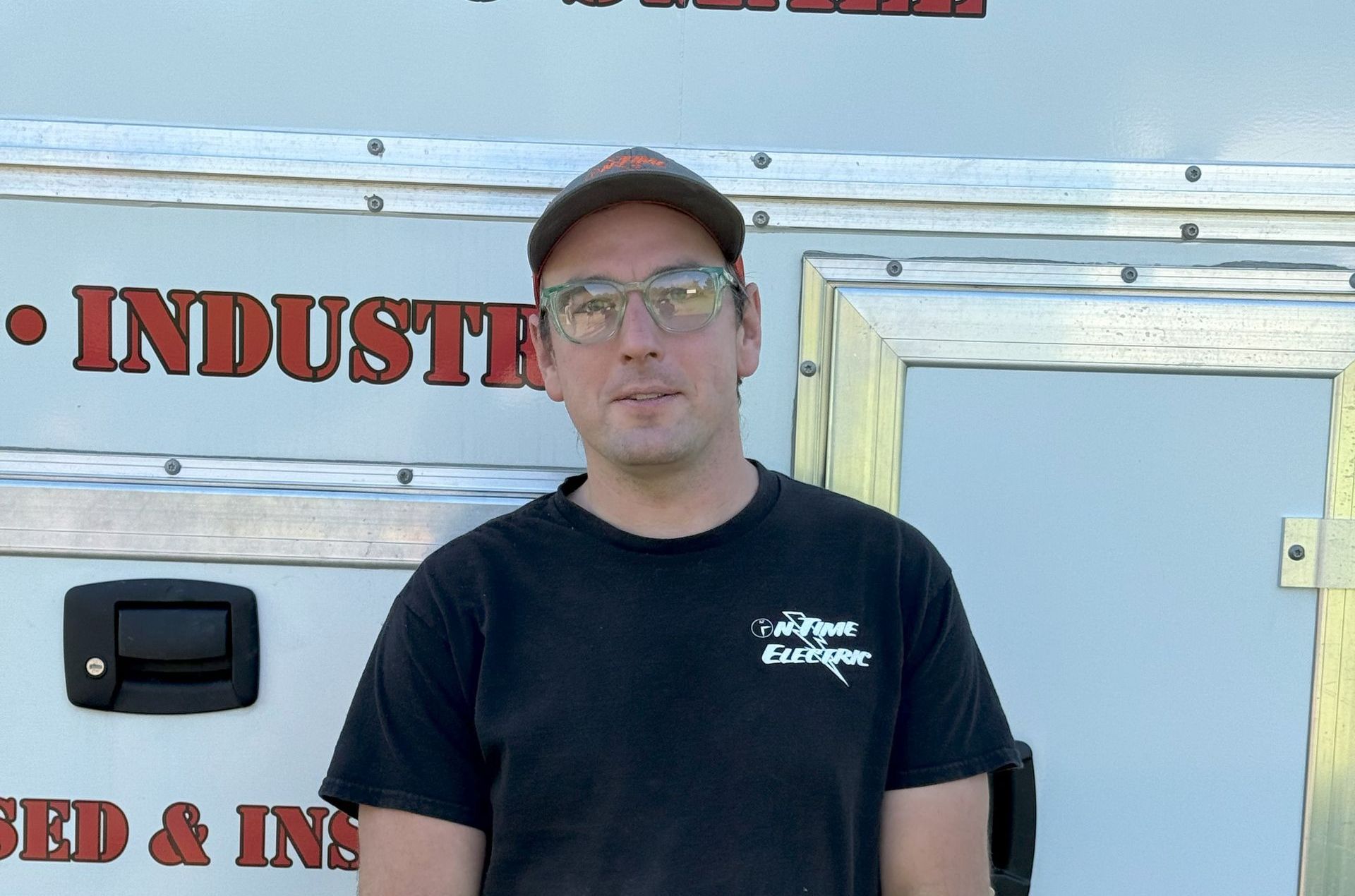 A man wearing glasses and a hat is standing in front of a trailer that says induste