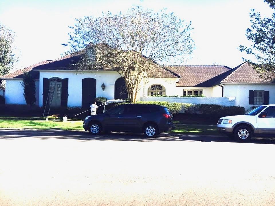 White house with brown roof, blue SUV and white truck parked on the street. Person near the house is painting.