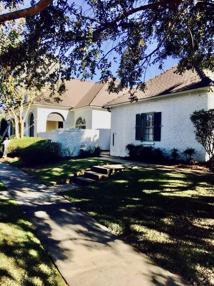 White stucco house with dark shutters, brick roof, and arched entryway; shaded front yard.