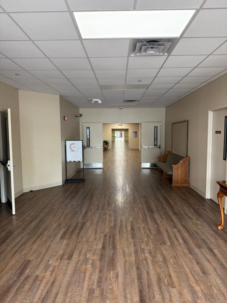 Long hallway with wood-look floor, beige walls, and white ceiling with recessed lighting. Doors and seating along the walls.