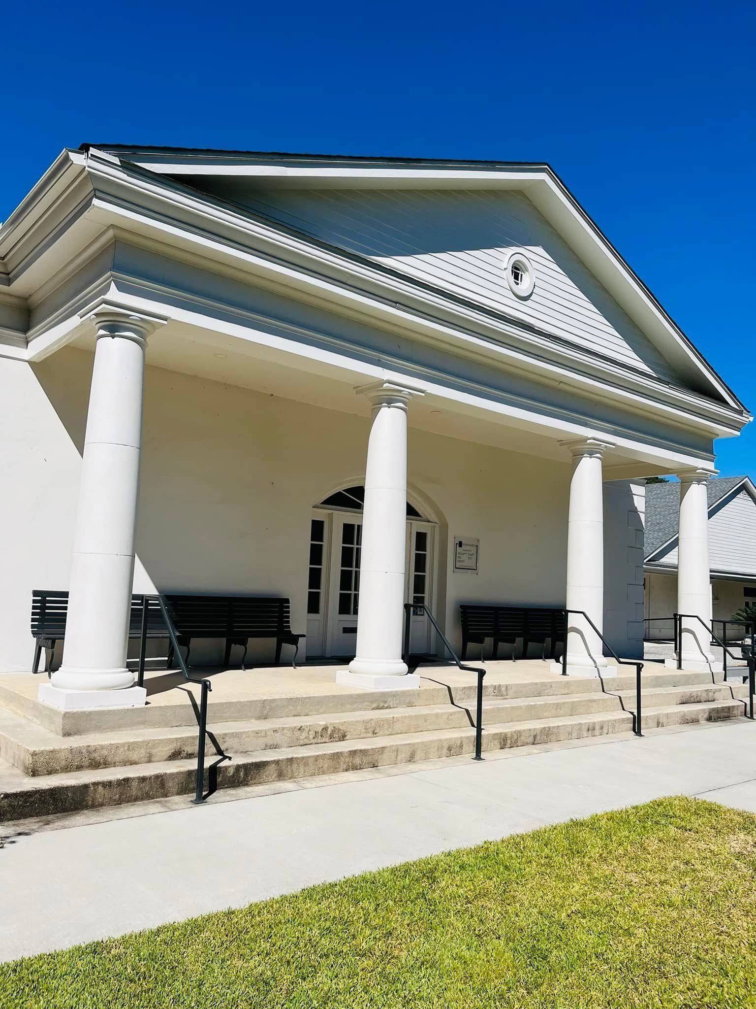 White building with columns, steps, and benches under a porch, set against a bright blue sky.