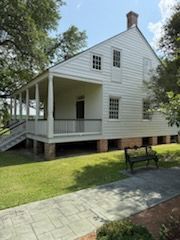 White two-story house with porch and brick foundation; walkway and bench in front, trees surround it.