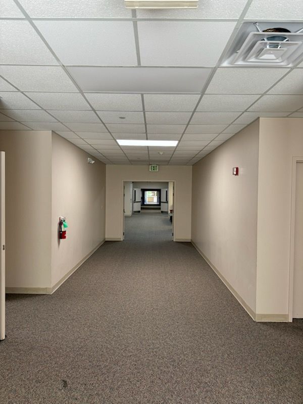 Hallway with beige walls, gray carpet, and a drop ceiling. A fire extinguisher and exit sign are visible.