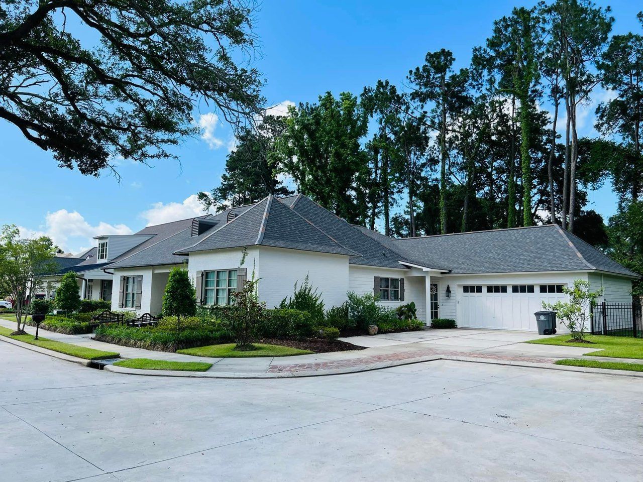 White house with gray roof on a corner lot, surrounded by greenery and trees under a blue sky.