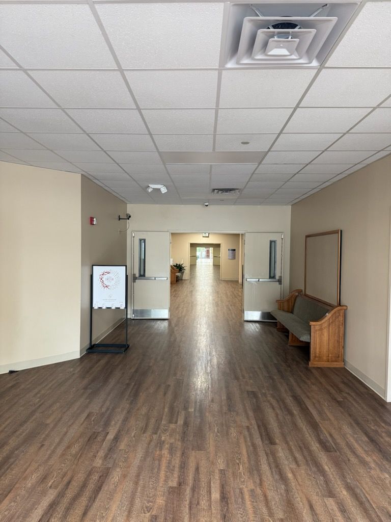 Hallway with brown wooden floors, beige walls, and white ceiling with recessed lighting. A bench and sign are visible.