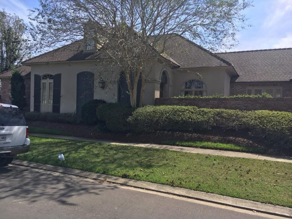 House with brown roof, stucco walls, shutters, and a well-manicured lawn and bushes.