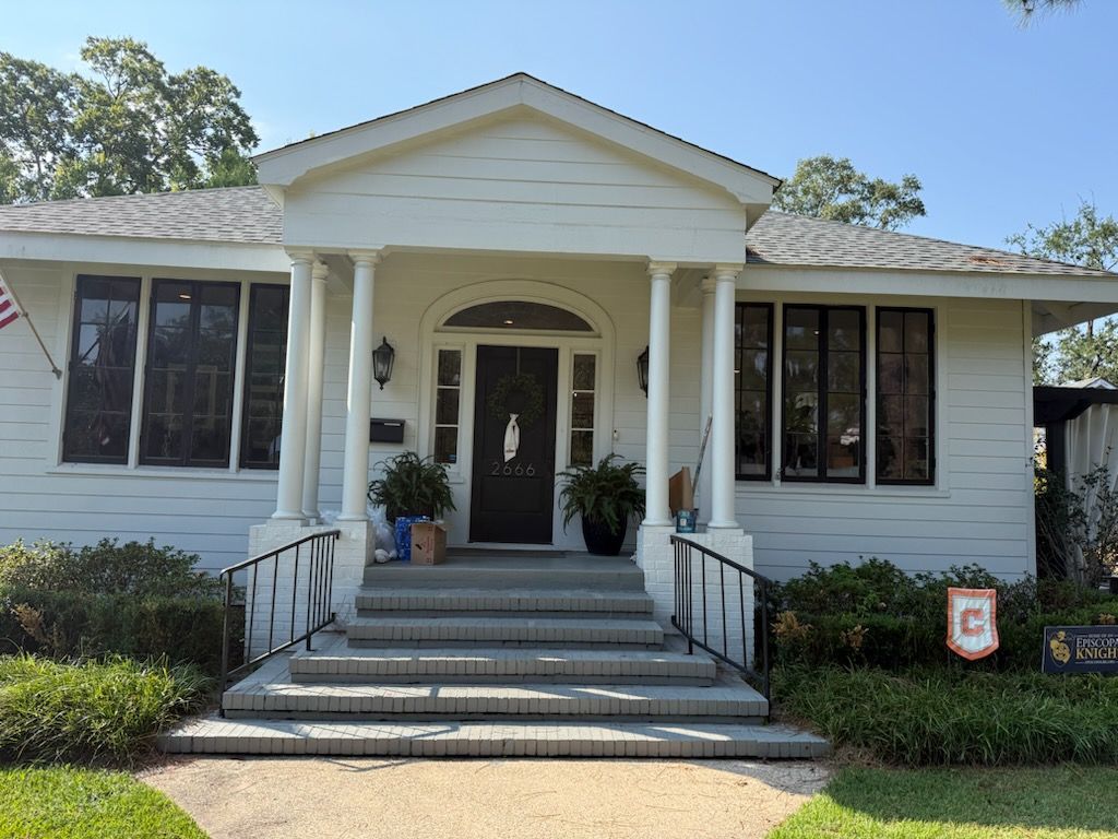 White house with porch columns and steps. Black windows, black door, and foliage.