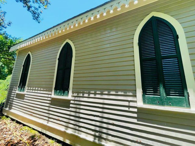Side view of a light brown wooden church with three arched windows and dark green shutters on a sunny day.