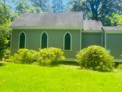 Green wooden church with three arched windows, green grass, and bushes outside.