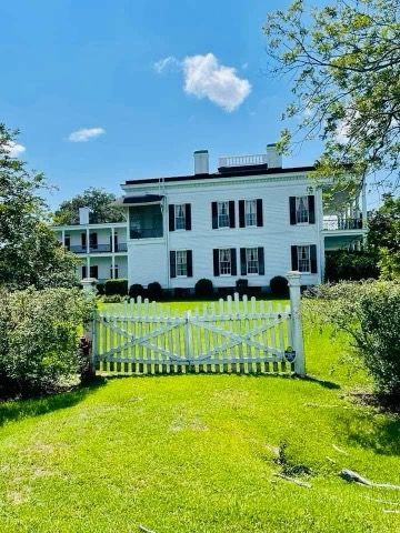 White house with black shutters, green lawn, white picket fence, blue sky.