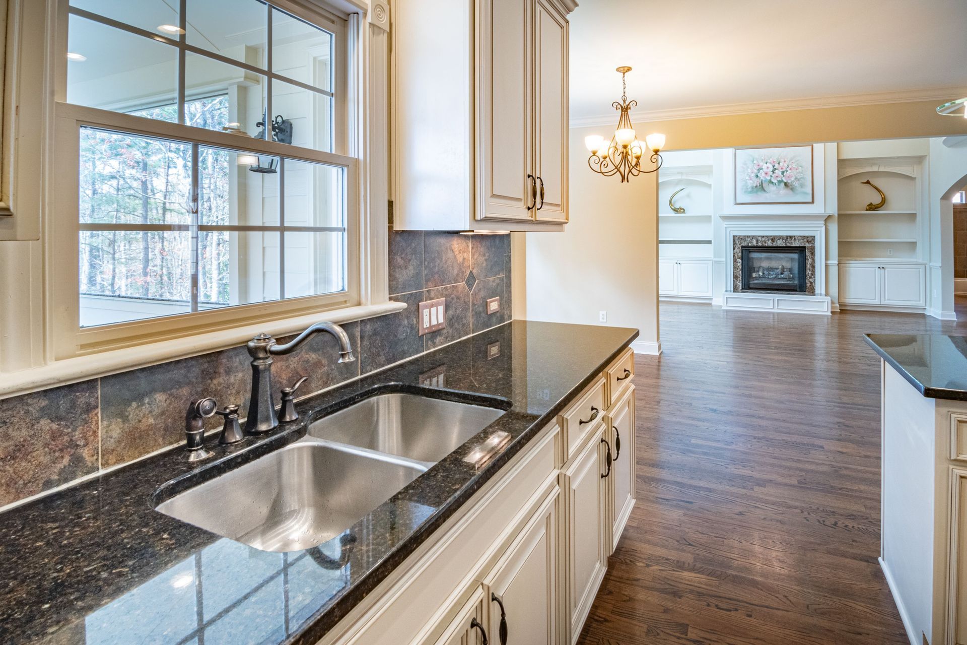 Bright kitchen with granite sink and white cabinets, opening to a living room with fireplace and chandelier