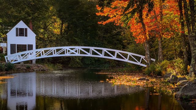 White covered bridge by a pond beside a white house in autumn woods