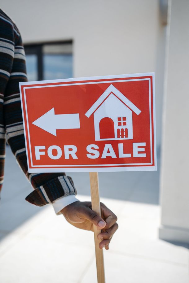 Hand holding a red “FOR SALE” sign with a white house icon and left arrow outdoors