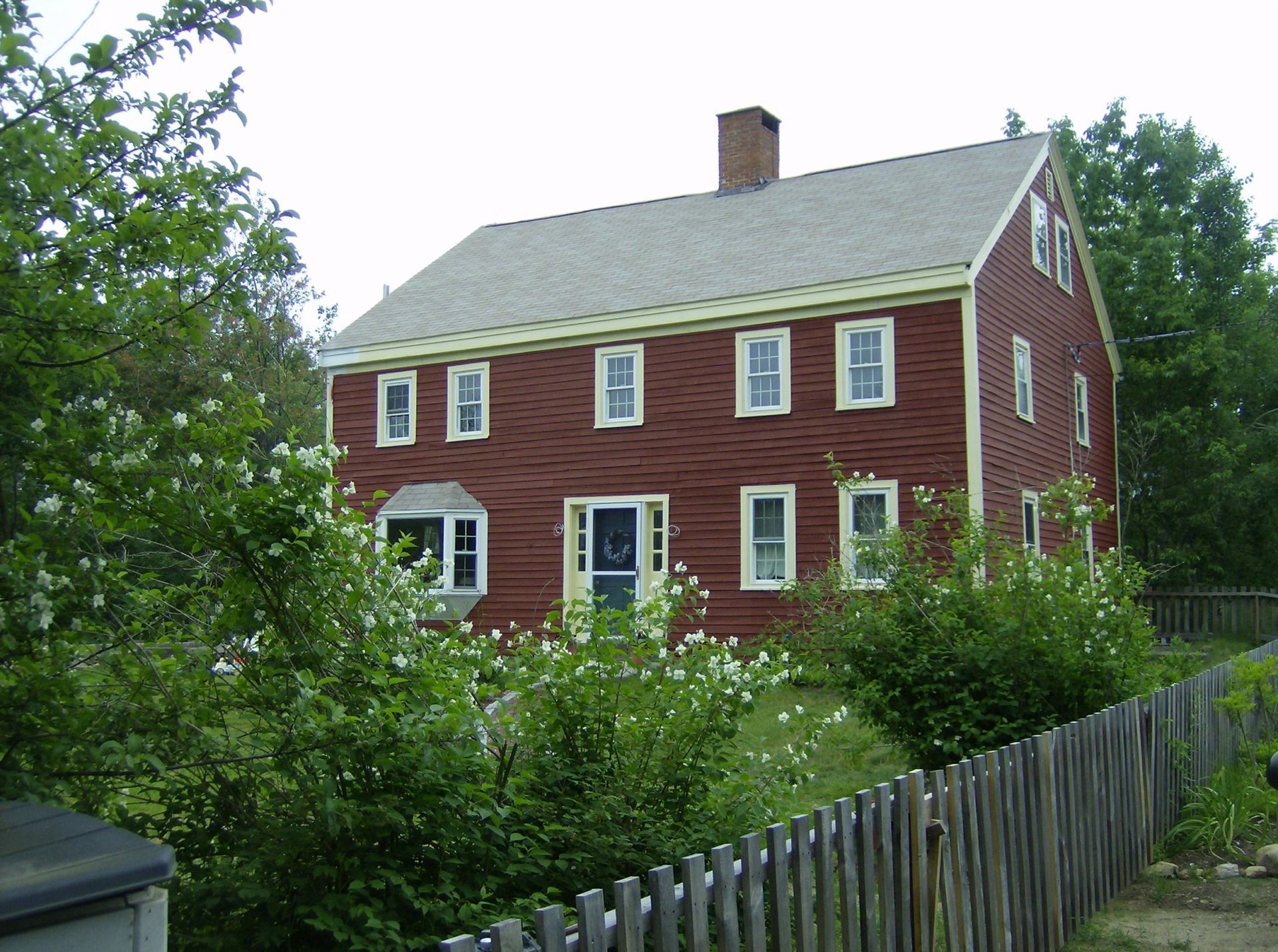 Red two-story house with white-trim windows, surrounded by greenery and a wooden fence.