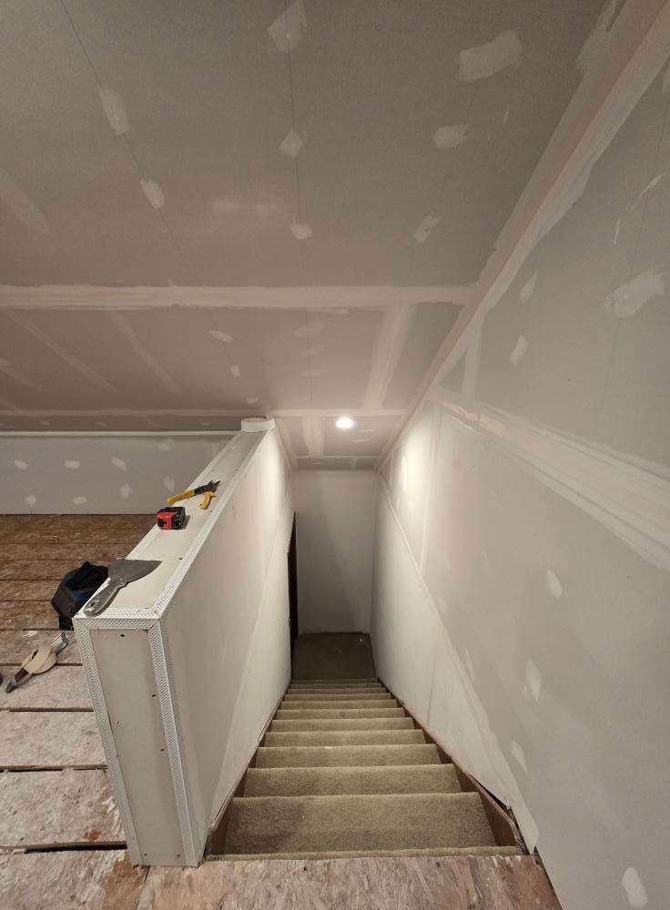 Stairway leading down with drywall installed on walls and ceiling. Drywall is mudded.