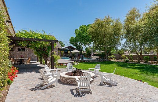 Stone patio with white chairs around a fire pit, overlooking a backyard with a pool and green lawn.