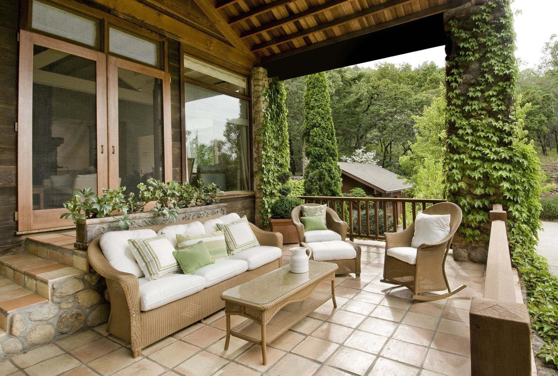 Patio with wicker furniture, stone steps, and climbing ivy-covered column. Lush greenery in background.