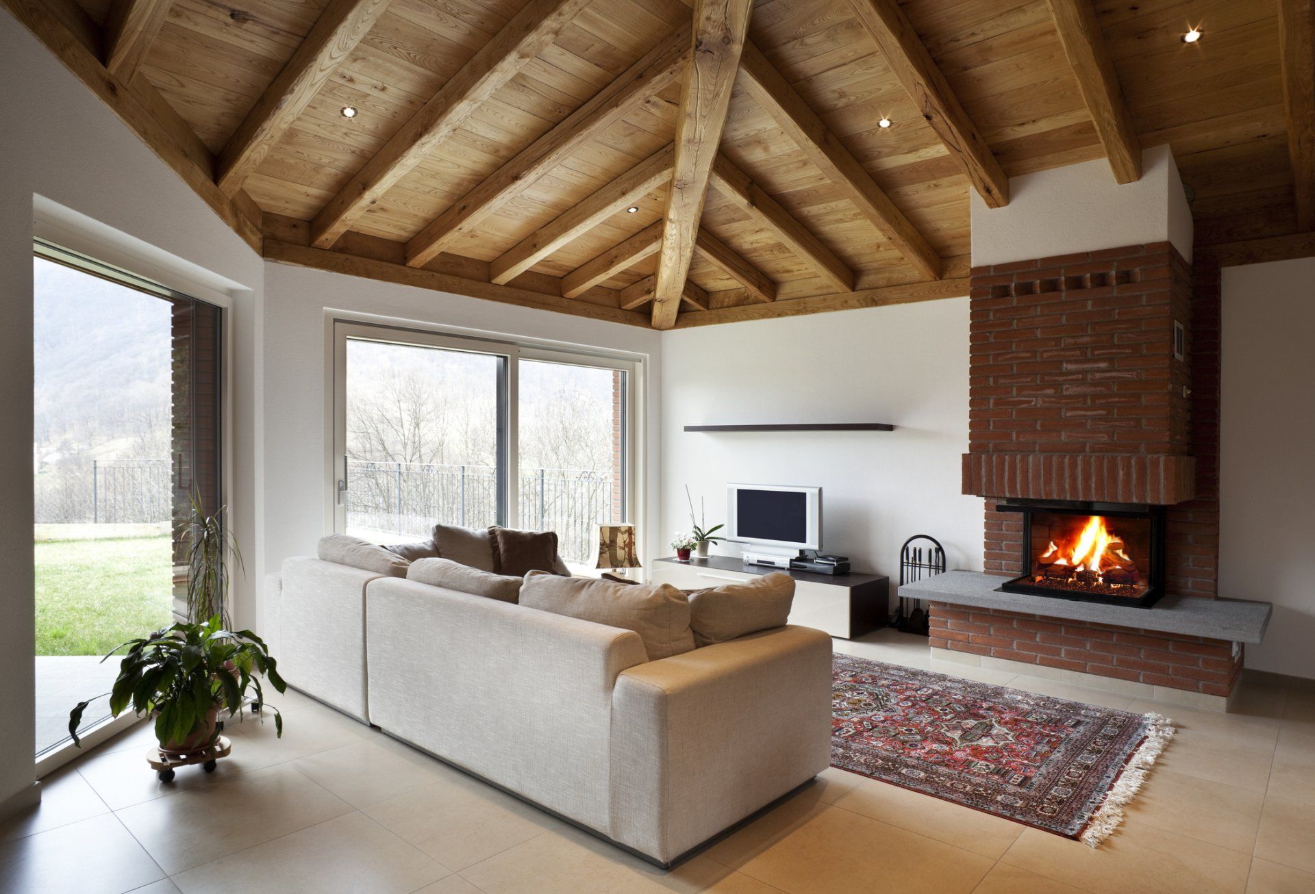 Living room with a brick fireplace, wooden ceiling, large windows, and a beige sofa.