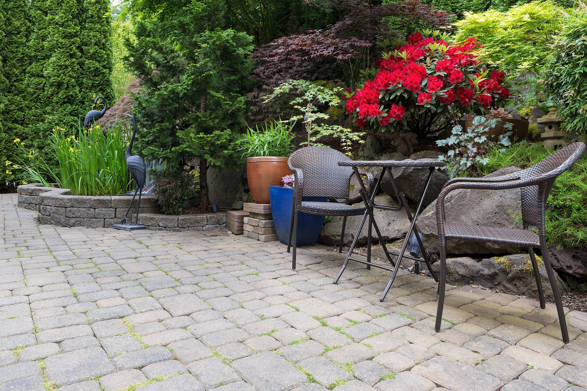 Patio with brick pavers, bistro set, surrounded by lush greenery and colorful flowers.