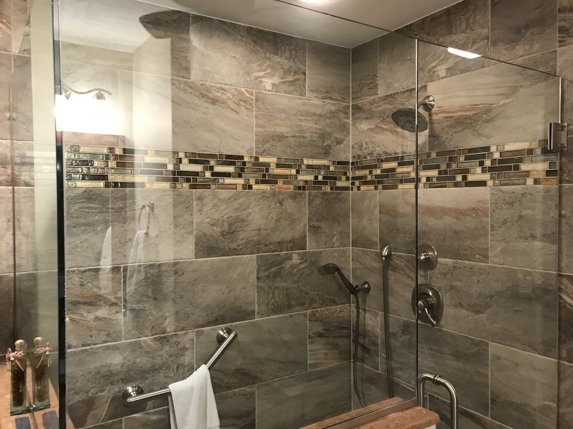 Shower with gray tile walls, glass door, and decorative band of mosaic tiles.