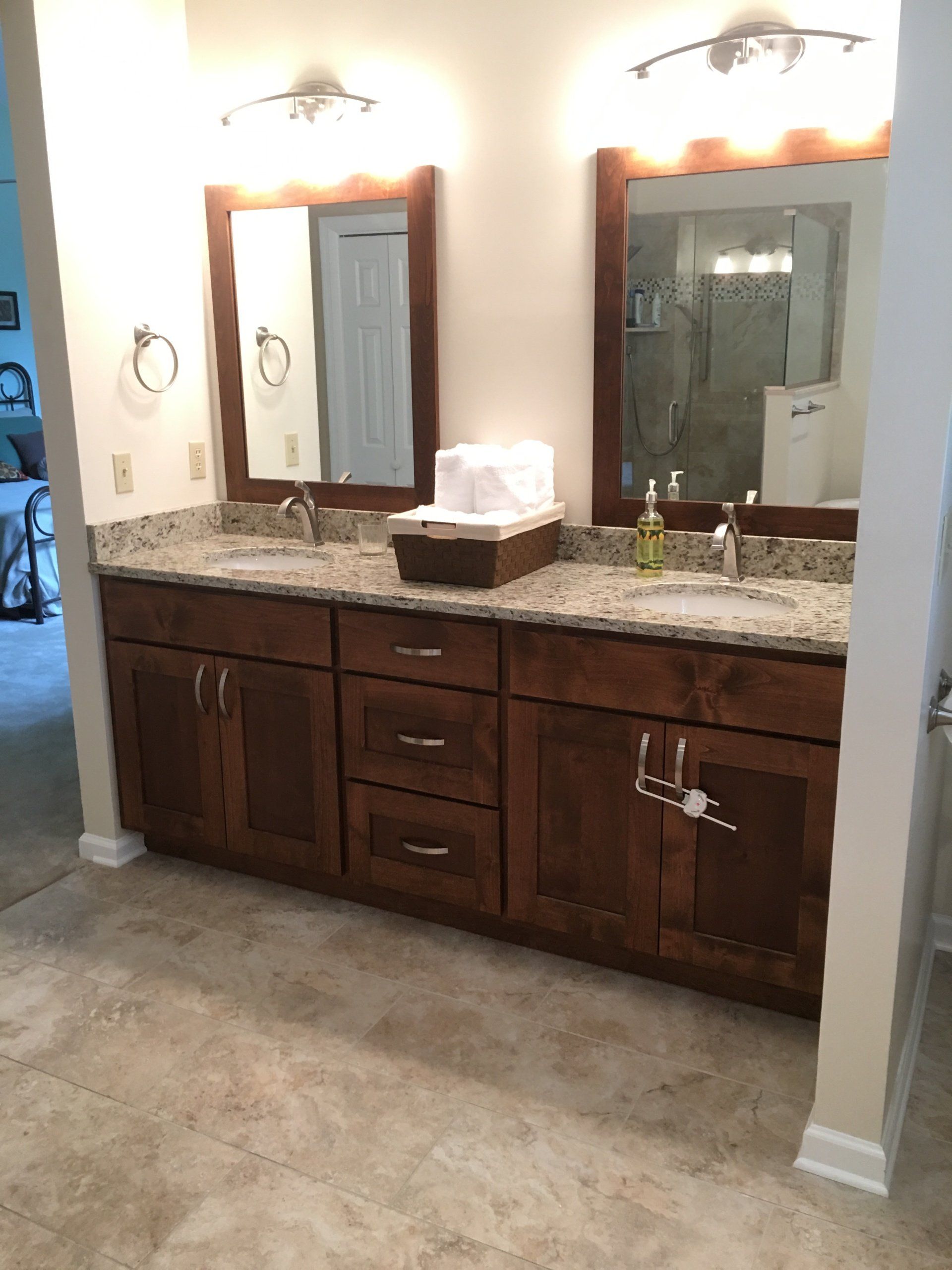 Bathroom with double sinks, brown cabinets, granite countertop, mirrors, and sconce lights.