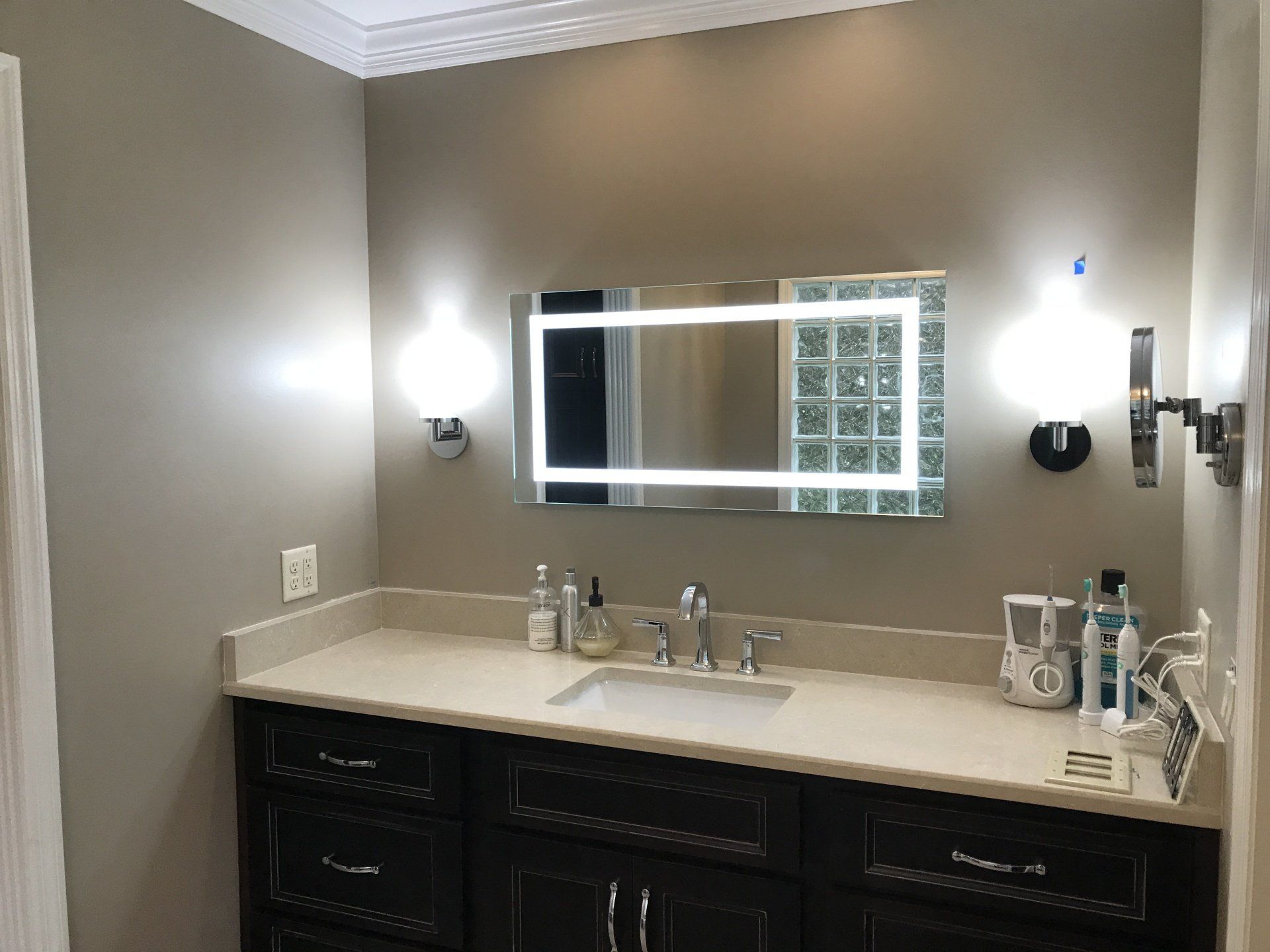 Bathroom with lit mirror, vanity, and wall sconces. Dark cabinets, light countertop, neutral walls.